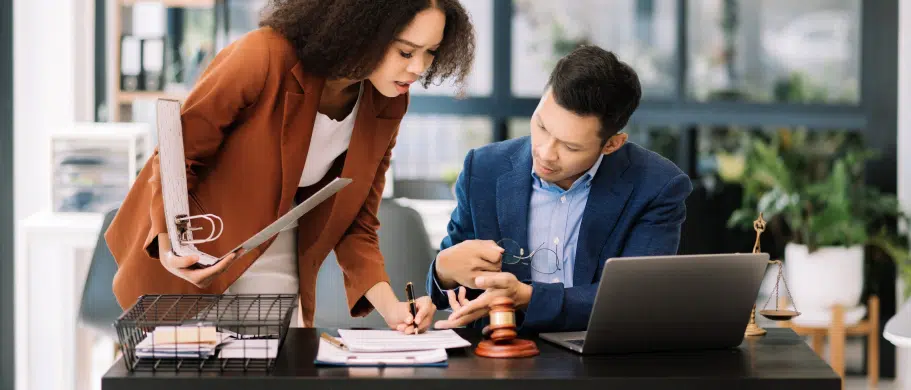 The image shows a female and a male lawyer in an office, engaged in a discussion about a case. They are seated at a desk, possibly reviewing documents or sharing insights.